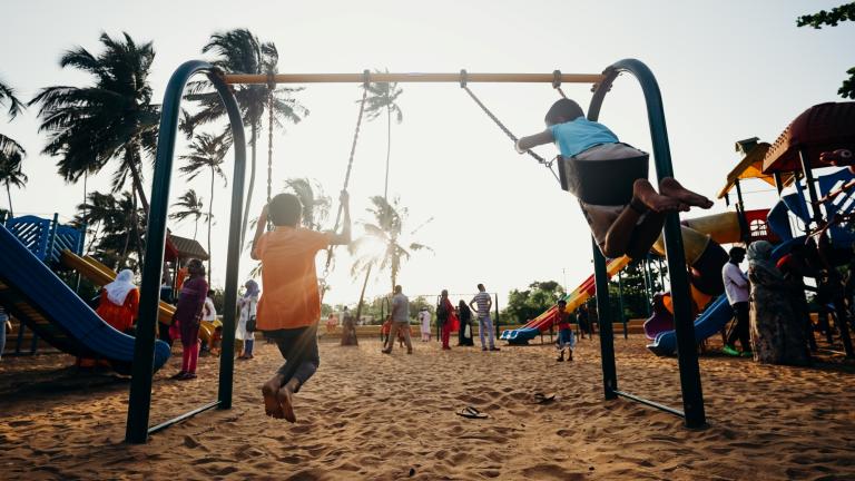 Crianças brincando em balanços em um parque de bairro repleto de árvores