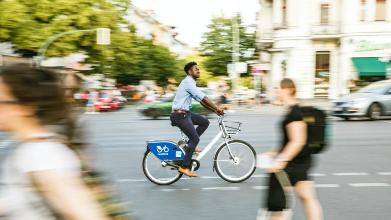 Uma imagem de uma cena de rua com pedestres e um ciclista. As árvores são vistas ao fundo.